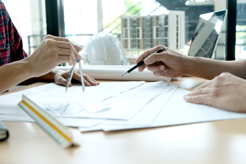 Zwei Personen arbeiten mit Zirkel und Stift an Architekturzeichnungen; im Hintergrund sind ein Laptop, weiße Bauhelme und ein Modell eines Gebäudes zu sehen.
