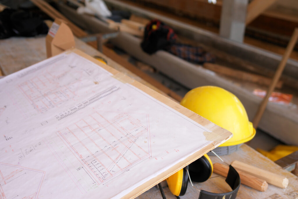 Technische Bauzeichnung auf einem Holztisch in einer Werkstatt; daneben ein gelber Schutzhelm und Gehörschutz in einer Baustellenumgebung.