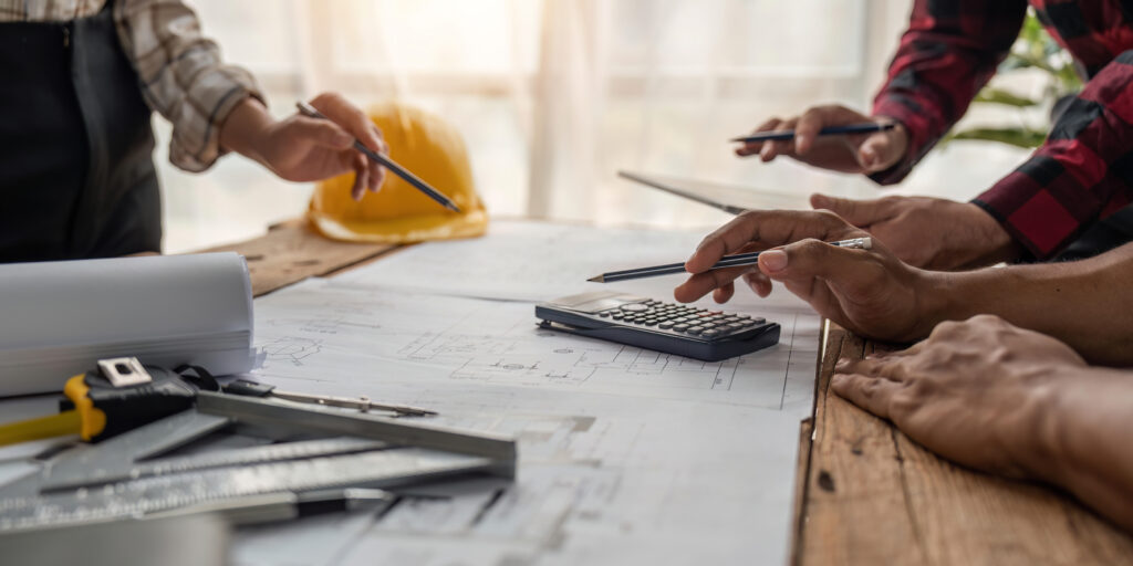 Architekten oder Bauingenieure diskutieren Baupläne am Tisch, mit einem Taschenrechner, Linealen und einem gelben Schutzhelm im Hintergrund.