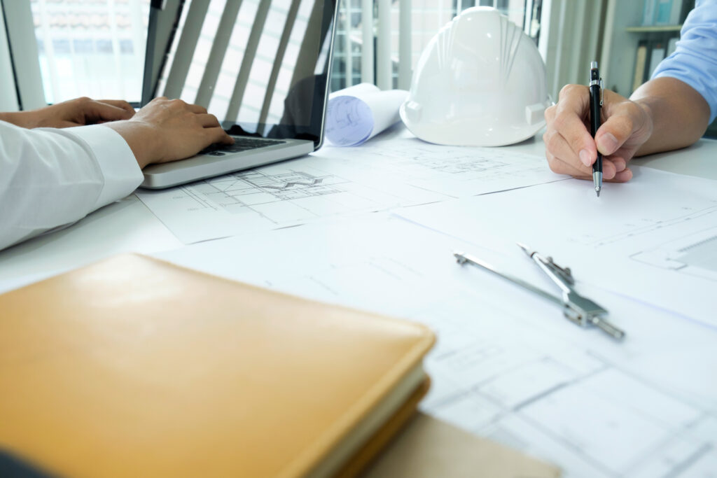 Zwei Personen arbeiten an einem Architekturbüro-Tisch: Eine schreibt auf Bauplänen, die andere tippt am Laptop. Auf dem Tisch liegen ein weißer Schutzhelm, ein Zirkel, eine Mappe und technische Zeichnungen.