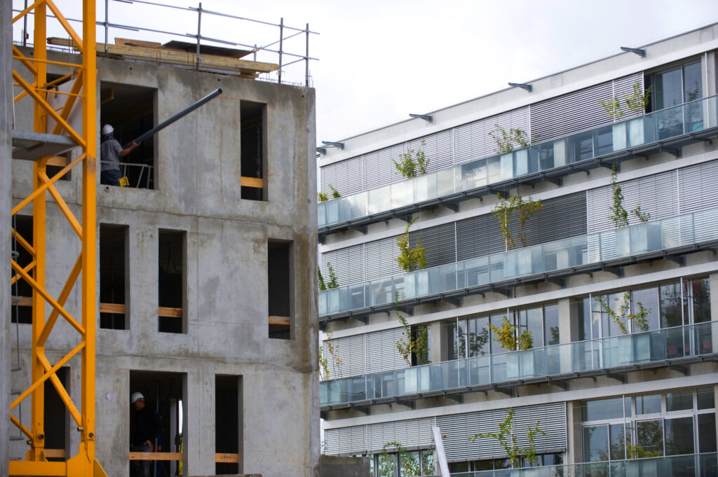 Bauarbeiter auf einer Baustelle eines Rohbaus mit gelbem Kran im Vordergrund; im Hintergrund ein modernes Wohngebäude mit Glasbalkonen und Bepflanzung.